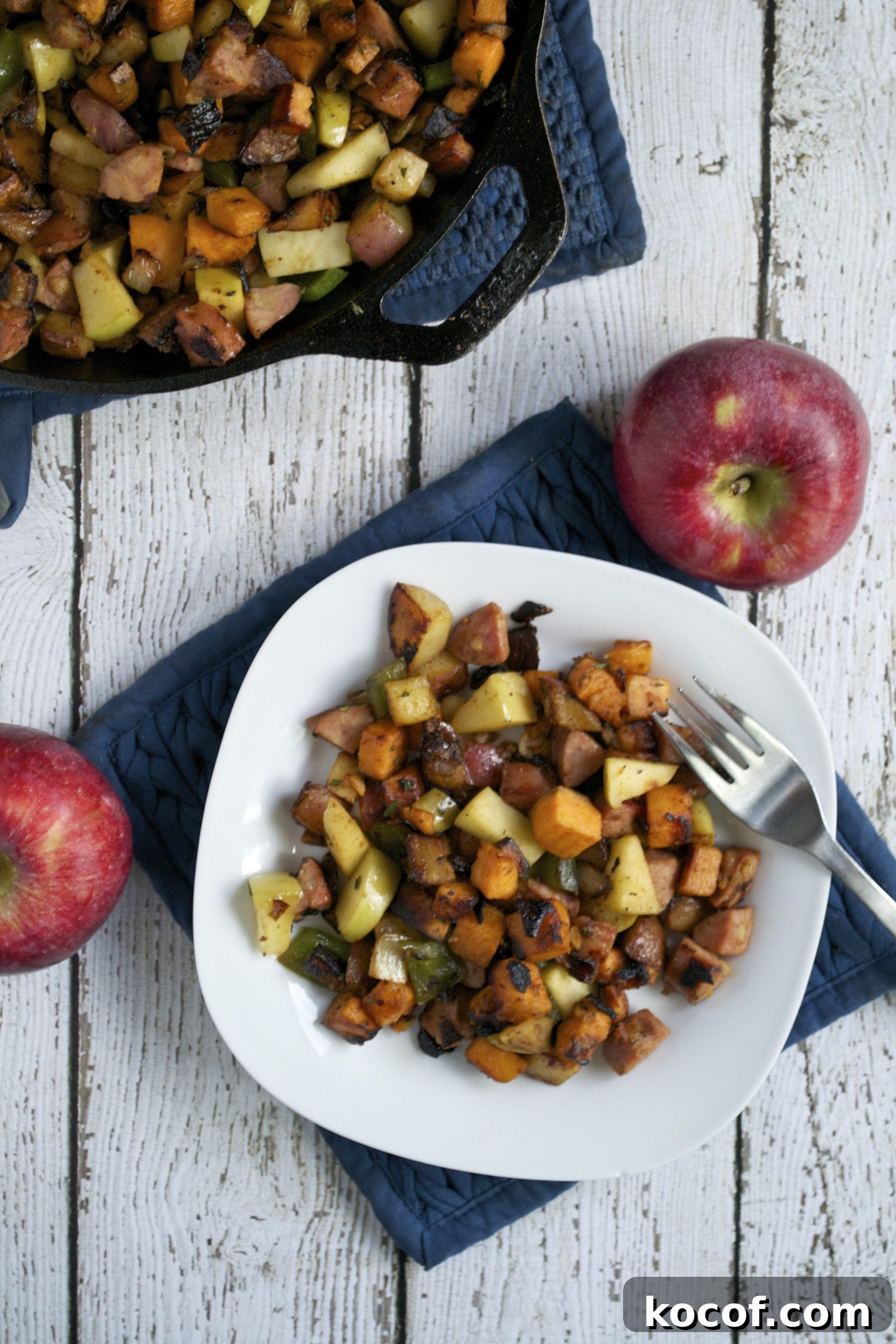Sweet Potato, Apple & Sausage Hash: Savory Breakfast Skillet 6 A white plate with Sweet Potato, Apple and Sausage Breakfast Hash sits on a blue quilted mat beside a fork. Two whole red apples and a skillet with more hash rest on a rustic white wooden table.