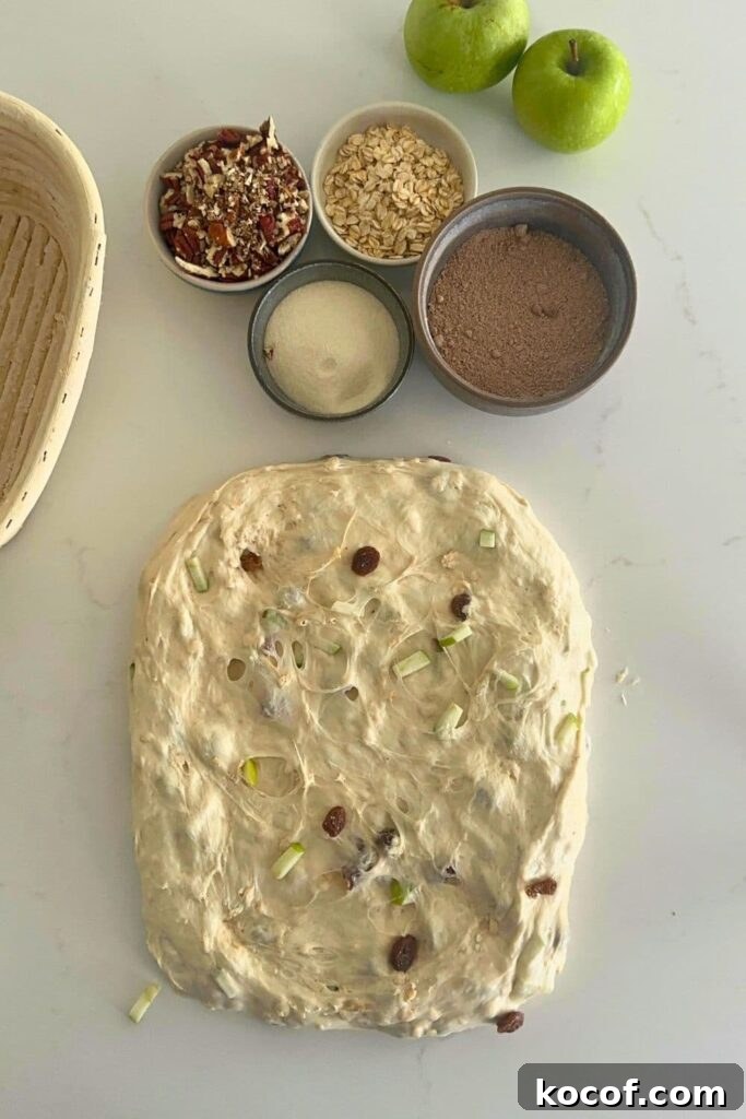A rough rectangle of dough that is ready to shape into a batard. There are 4 bowls of inclusions ready to sprinkle over the dough. 2 green apples can also be seen in the photo but these are just there for decoration.