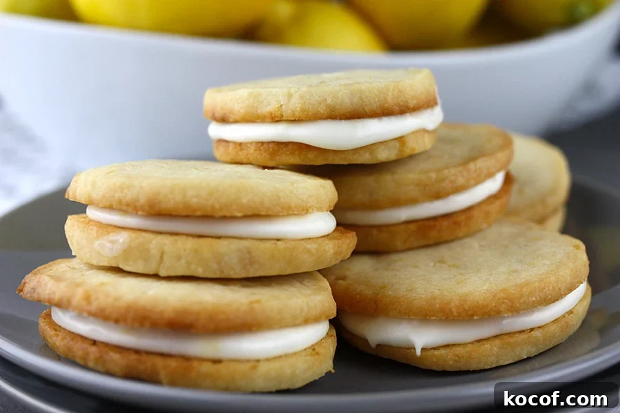 Lemon Sandwich Cookies stacked up on a plate with a bowl of lemons behind them.