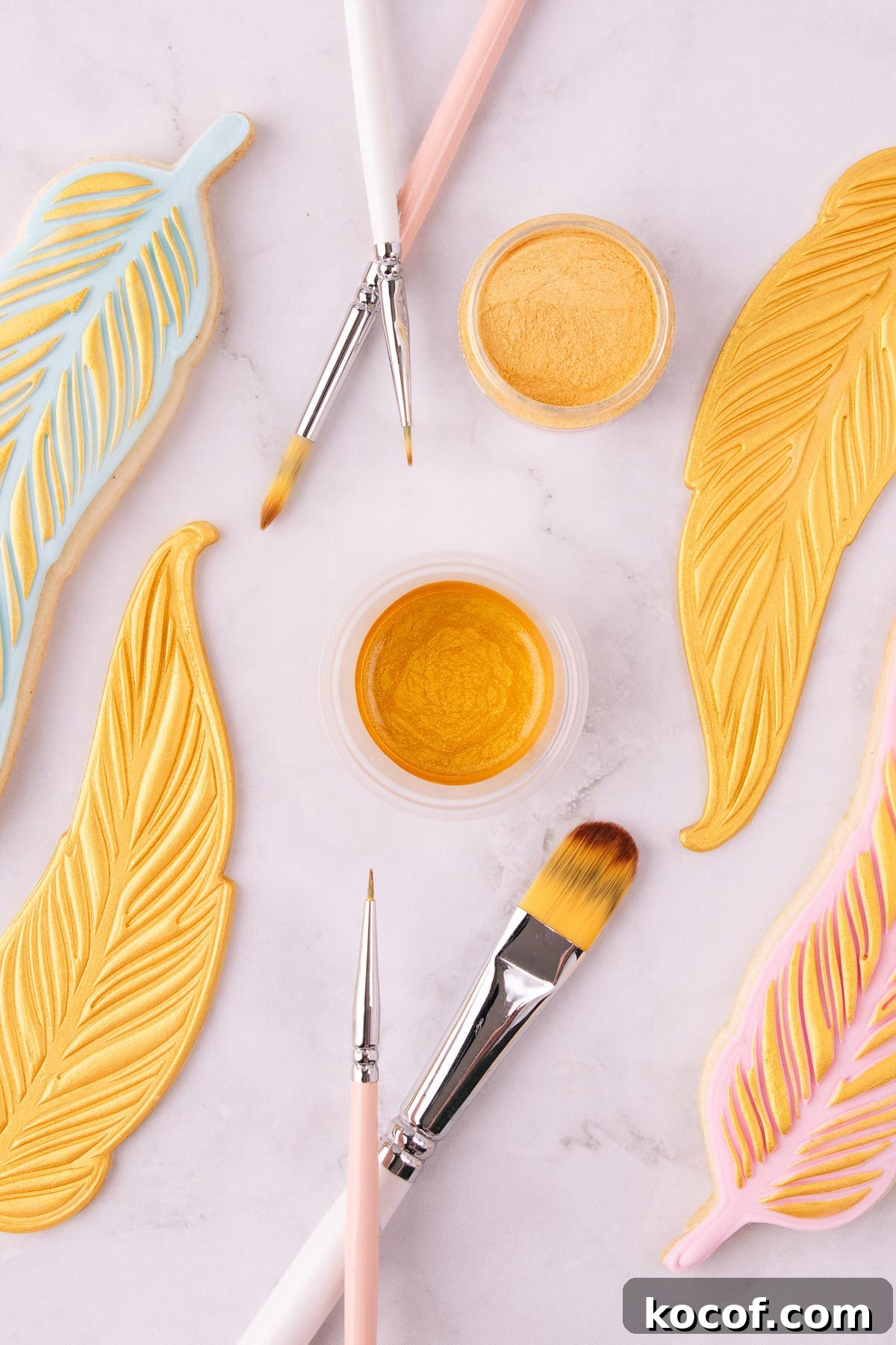 Make Edible Gold Paint at Home: A Step-by-Step Guide 2 A small plastic container of edible gold paint, surrounded by pink and white handled paintbrushes, and feather-shaped cookies painted with gold paint.