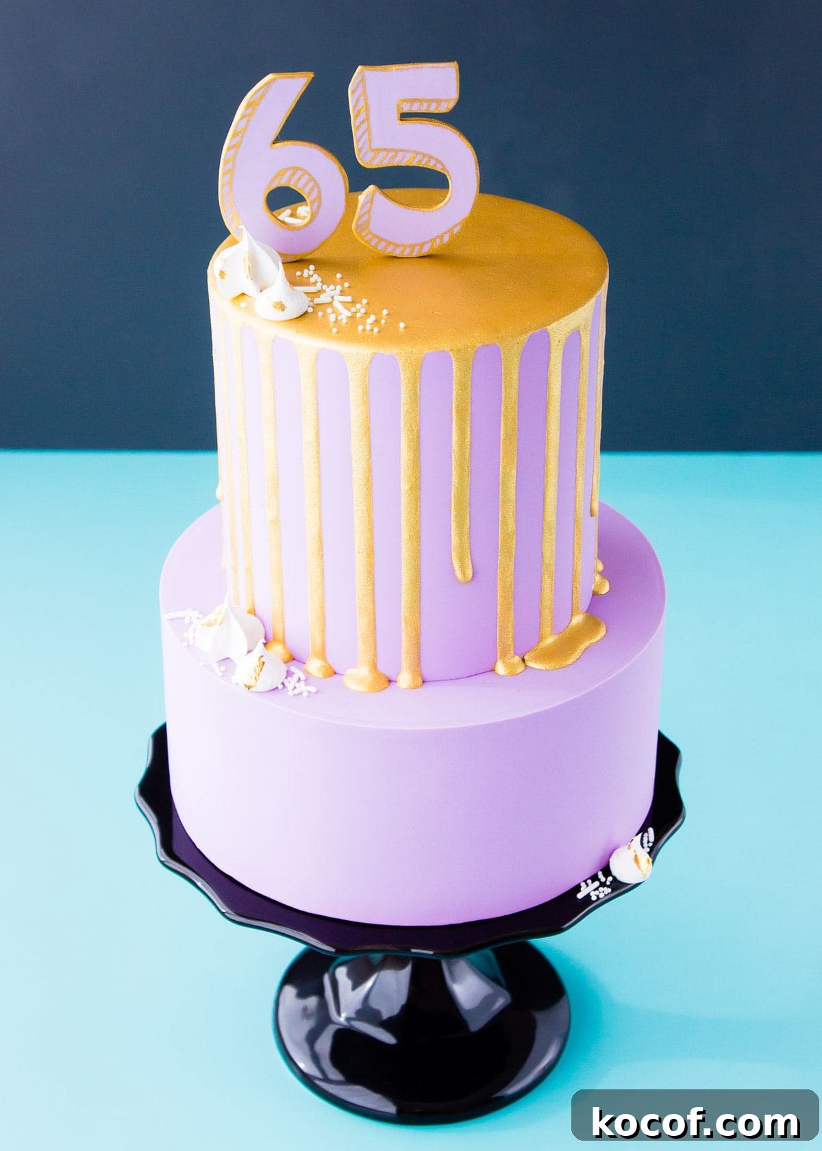 A two-tier cake covered in light purple fondant, with a gold drip on the top tier, a "65" number cake topper, and mini meringues, sitting on aa black cake stand.