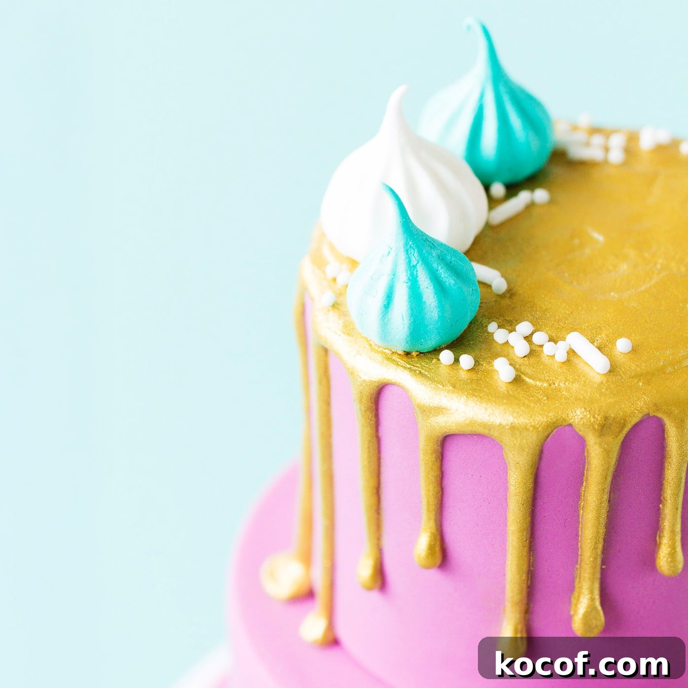 Close up of the top tier of a mini two tier cake covered with purple fondant icing with a metallic gold painted drip on the top tier, and mini meringues.