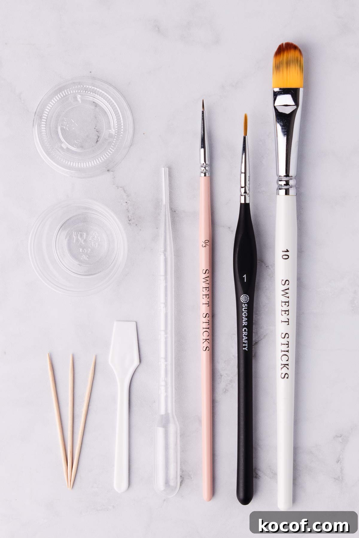 The tools and supplies used for making and using edible gold paint - paintbrushes, toothpicks, a clear plastic pipette and a small plastic container, on a light grey marble background.