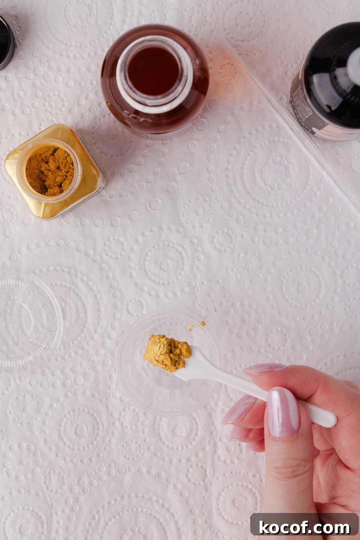 Make Edible Gold Paint at Home: A Step-by-Step Guide 9 A hand holding a small white spoon with about 1/4 teaspoon of gold edible lustre powder on the end, over a small plastic container.