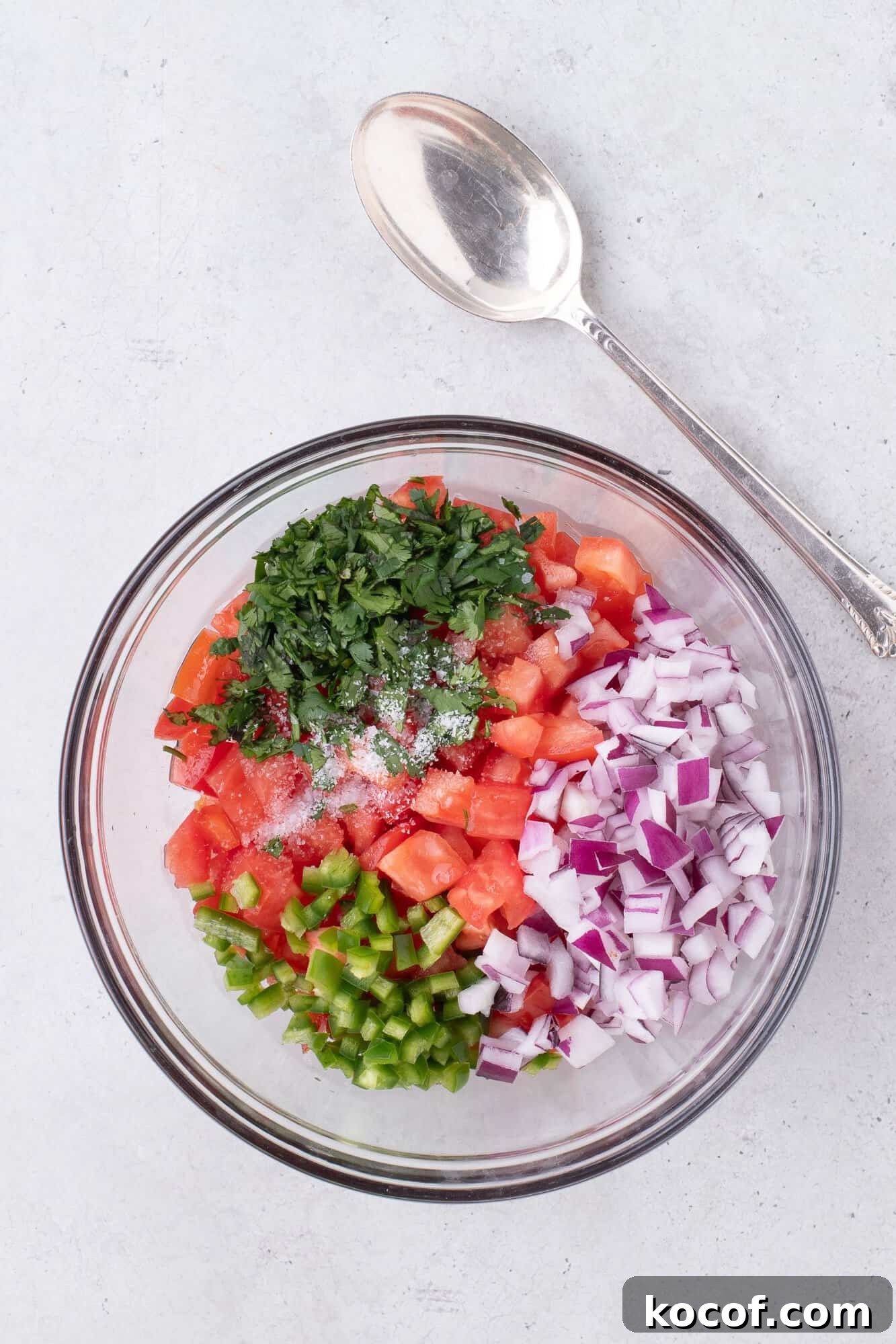A clear bowl with chopped tomatoes, red onions, green peppers, and cilantro, seasoned with salt, next to a silver spoon on a light gray surface.