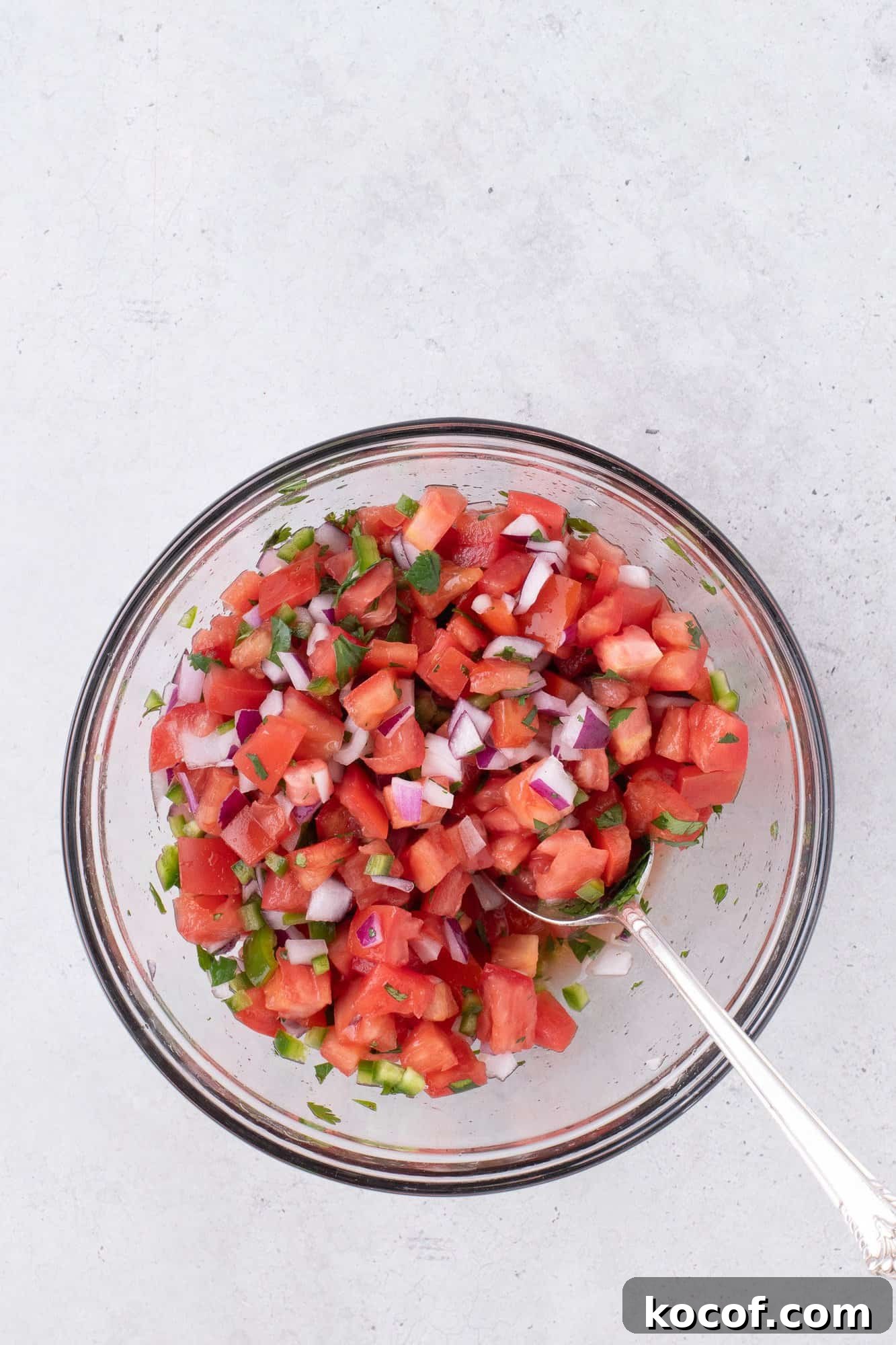 A clear bowl filled with fresh diced tomatoes, onions, jalapeños, cilantro, and a spoon on a white surface.