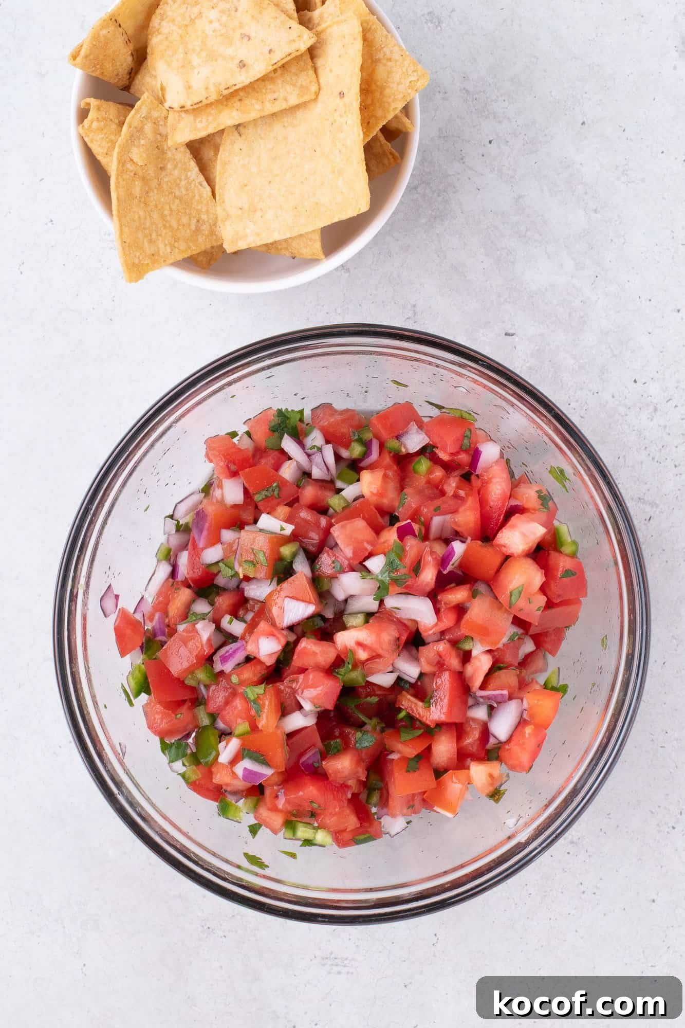 A bowl of pico de gallo with diced tomatoes, onions, and cilantro, next to a bowl of tortilla chips on a light surface.
