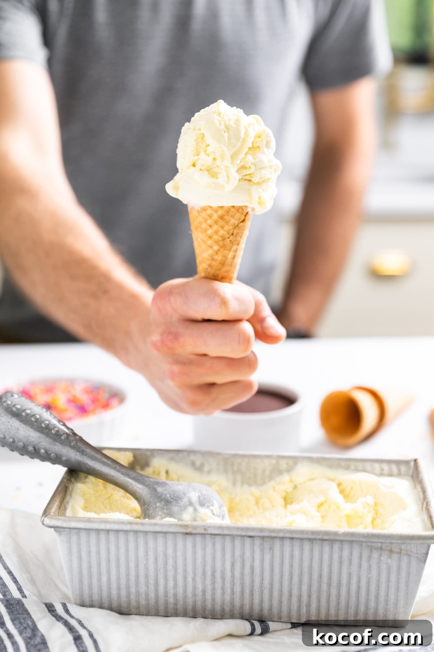 Hand holding ice cream cone filled with vanilla ice cream with rest of vanilla ice cream in silver container below sitting on countertop