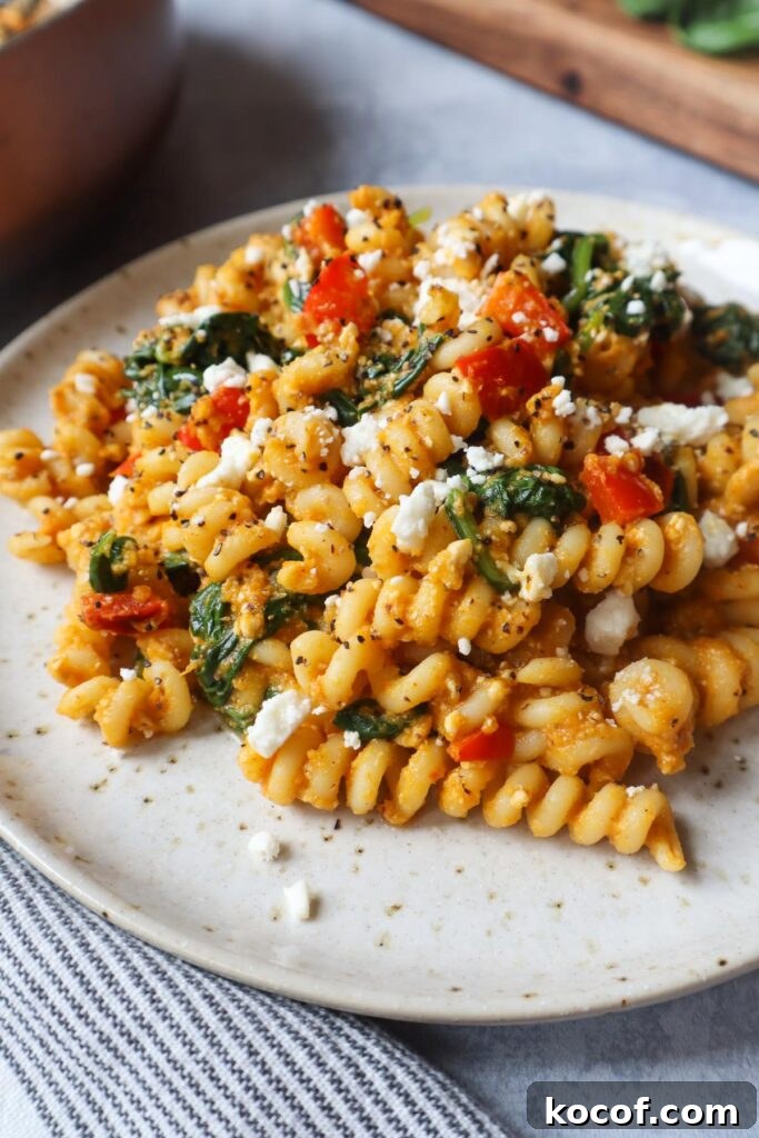 white plate with pasta tossed in a pumpkin feta sauce topped with red bell pepper and spinach