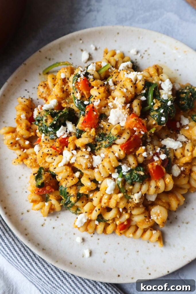 white plate with pasta tossed in a pumpkin feta sauce topped with red bell pepper and spinach