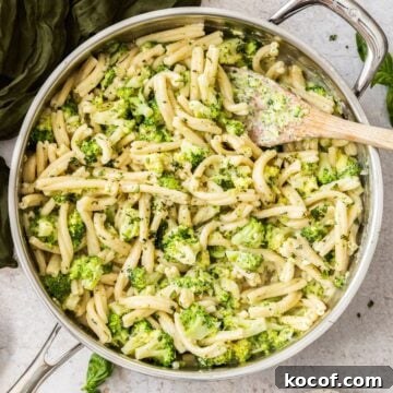 Large frying pan with Creamy Broccoli Pasta, sitting on a green cloth, with a wooden spoon in the pot.