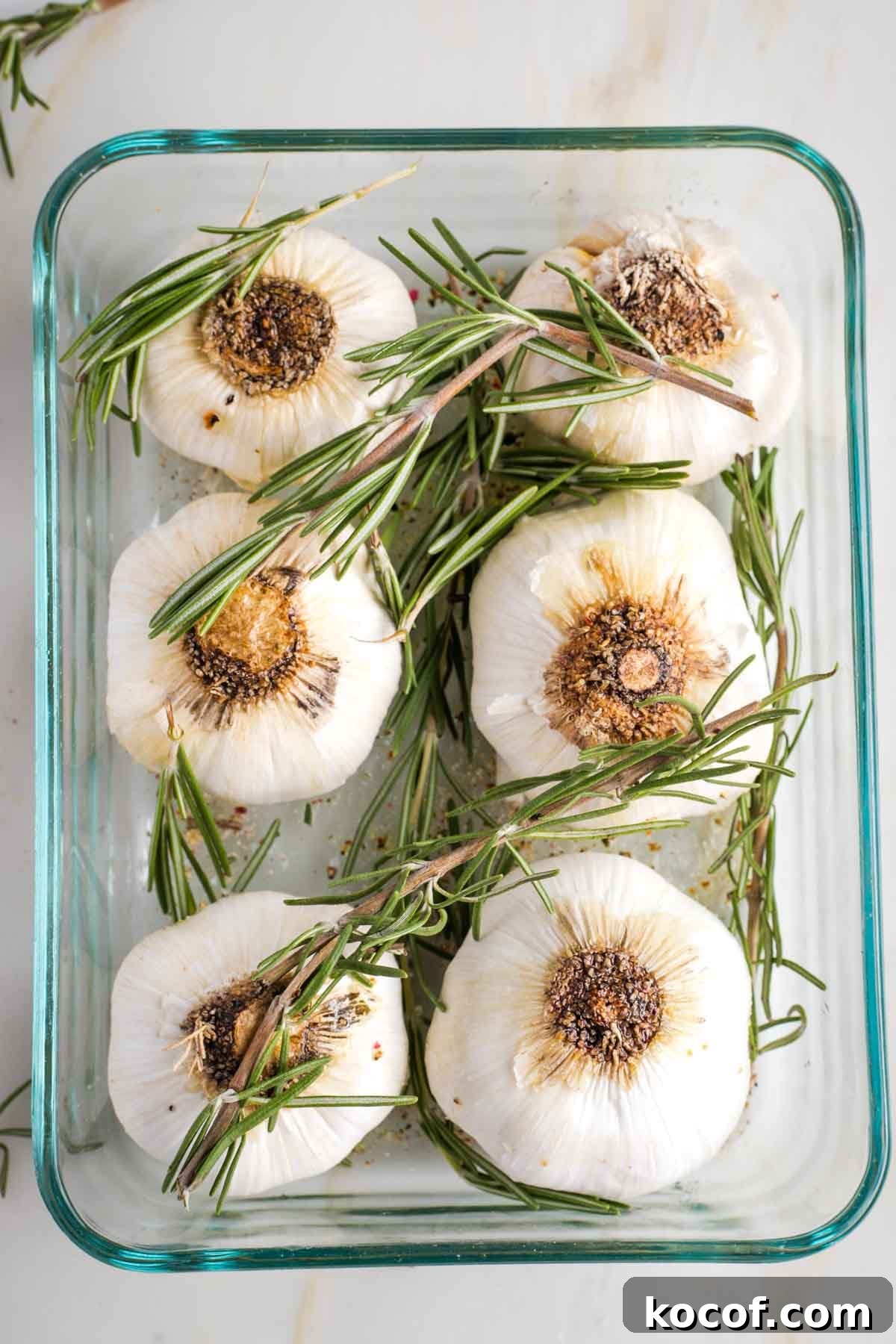 Oven-Roasted Garlic Bulbs: Crispy, Sweet Garlic Recipe 7 6 whole heads of garlic in a baking dish with fresh rosemary on top.