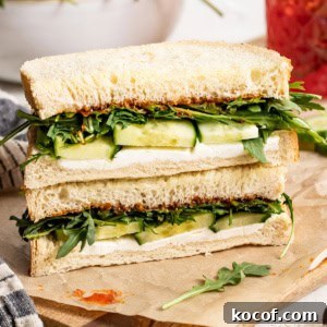 Two goat cheese sandwiches with cucumber and arugula stacked on top of each other on a cutting board.