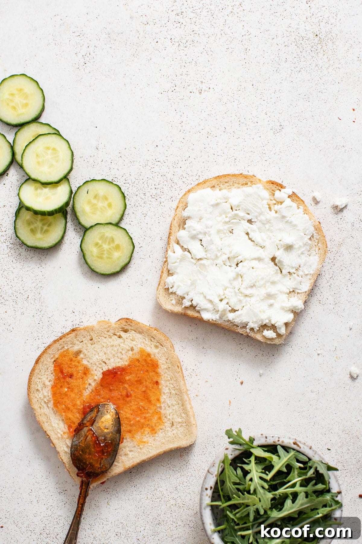 A spoon spreading jam on one side of the bread and goat cheese on the other.