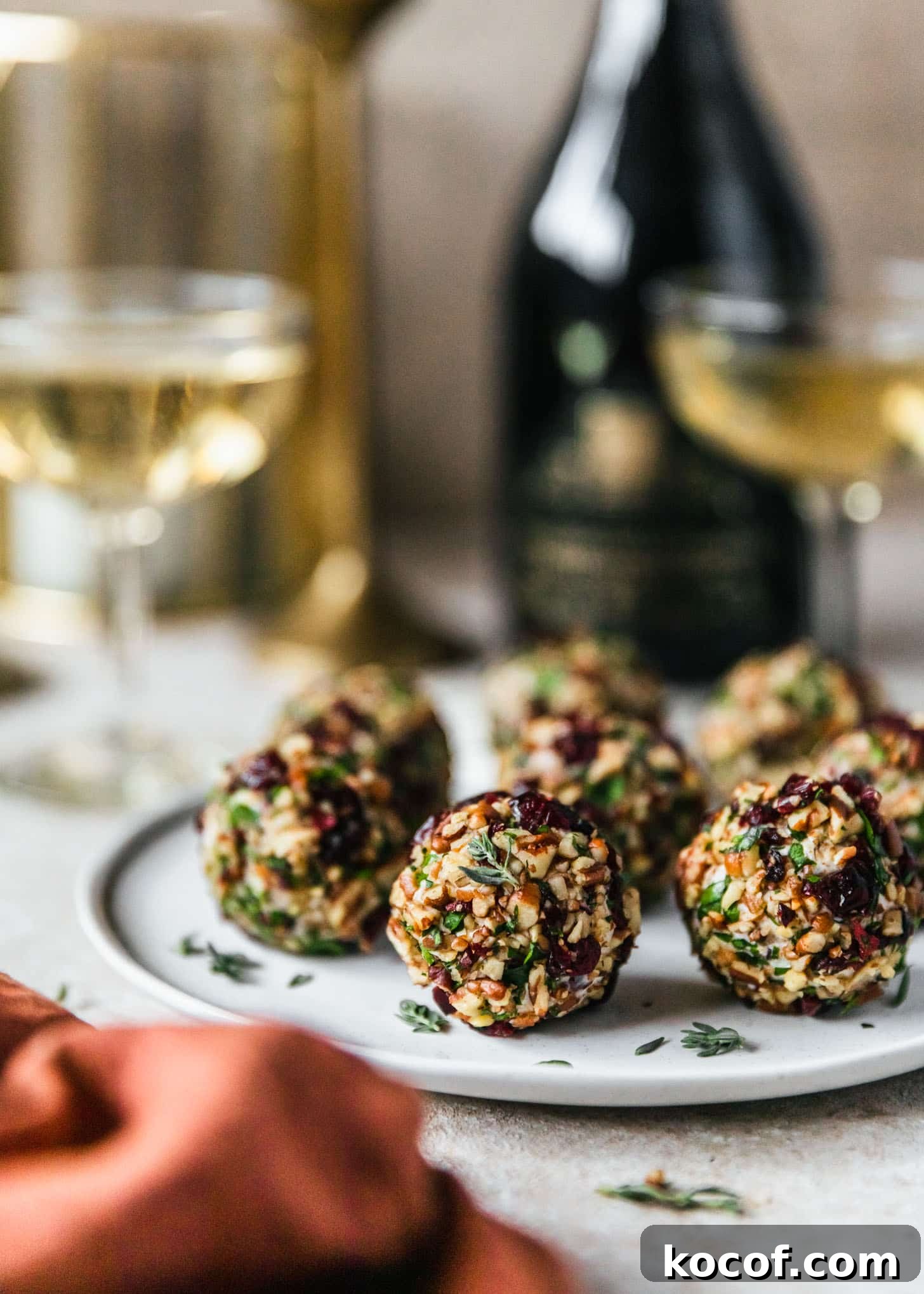 Cranberry Orange Cheesecake Bites: Festive Party Appetizer 2 Cranberry orange cheese balls on a white plate next to a bottle of champagne, glasses of champagne, and an orange napkin on a beige counter.