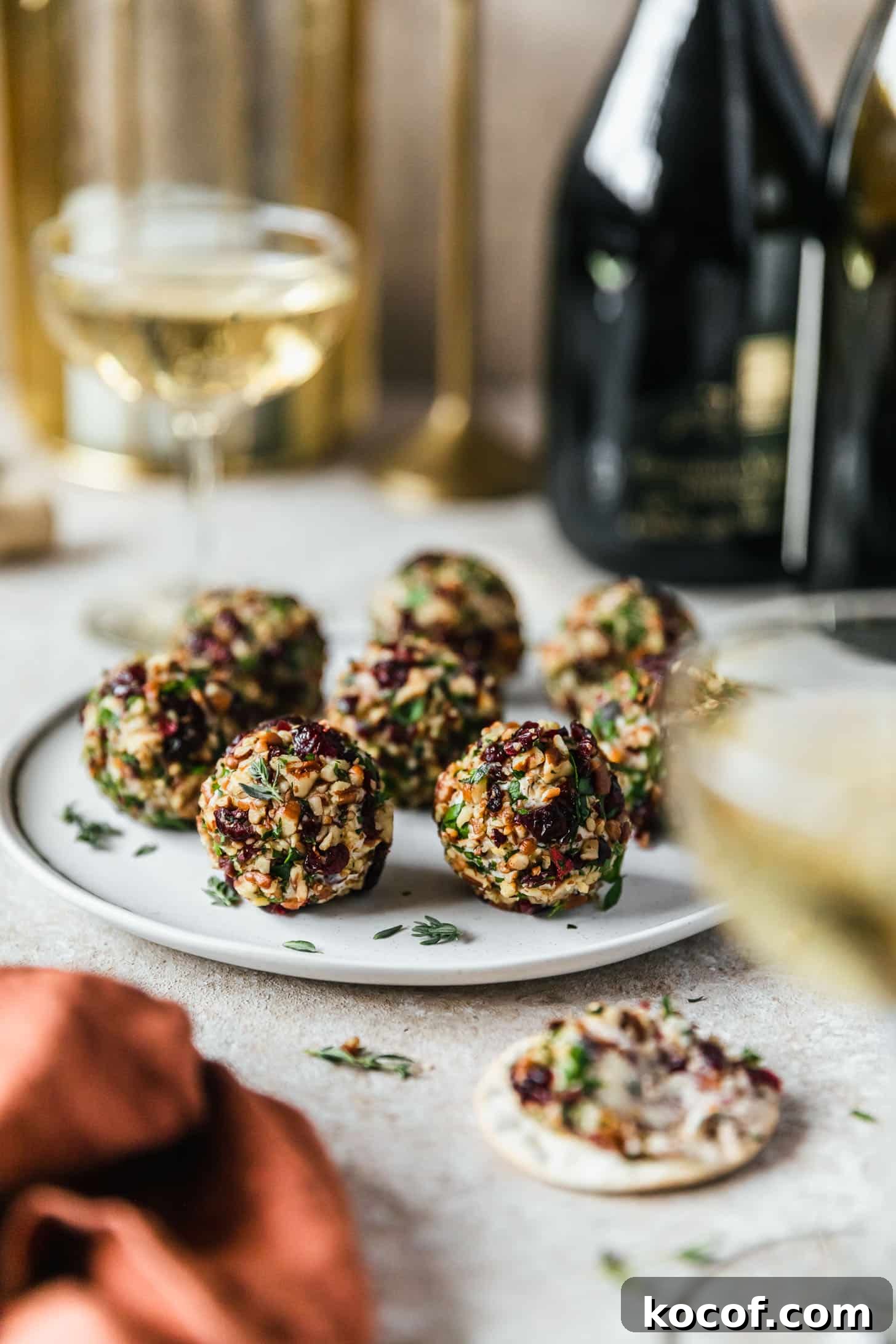 Cranberry Orange Cheesecake Bites: Festive Party Appetizer 11 A white plate of cranberry orange cheese ball bites on a beige counter next to a cracker, glasses of champagne, and a rust colored napkin.