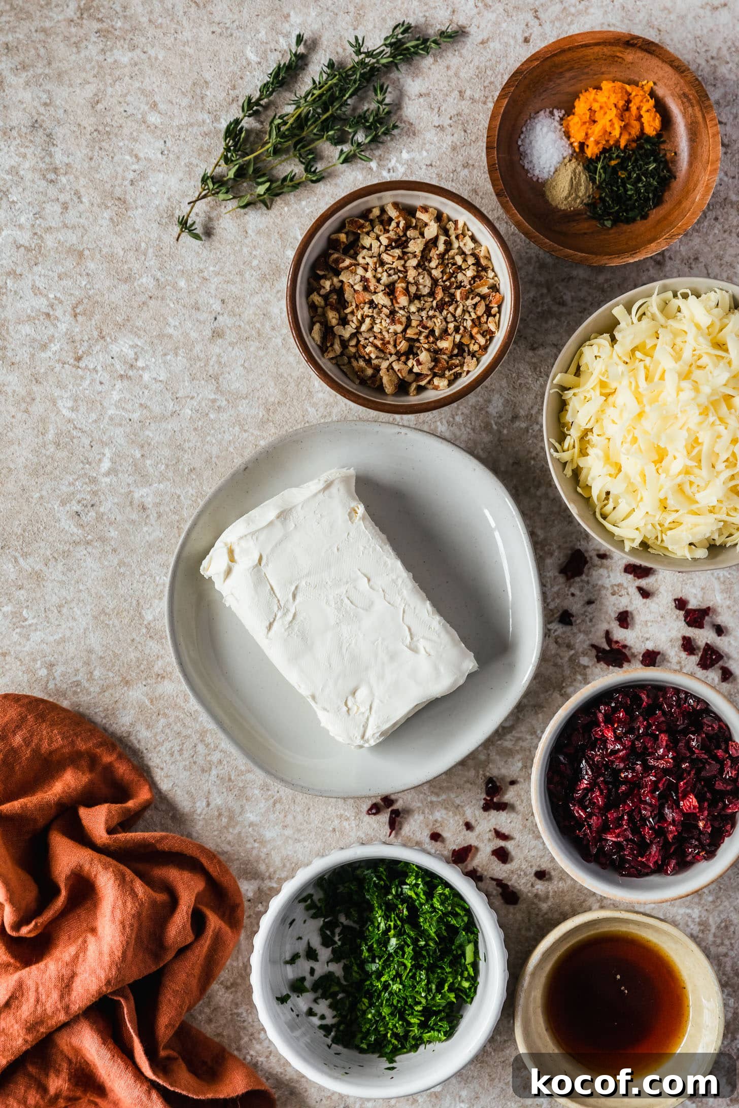 Cranberry Orange Cheesecake Bites: Festive Party Appetizer 3 A white plate of cream cheese on a tan counter next to white and brown bowls of cheese, pecans, dried cranberries, spices, and an orange linen.