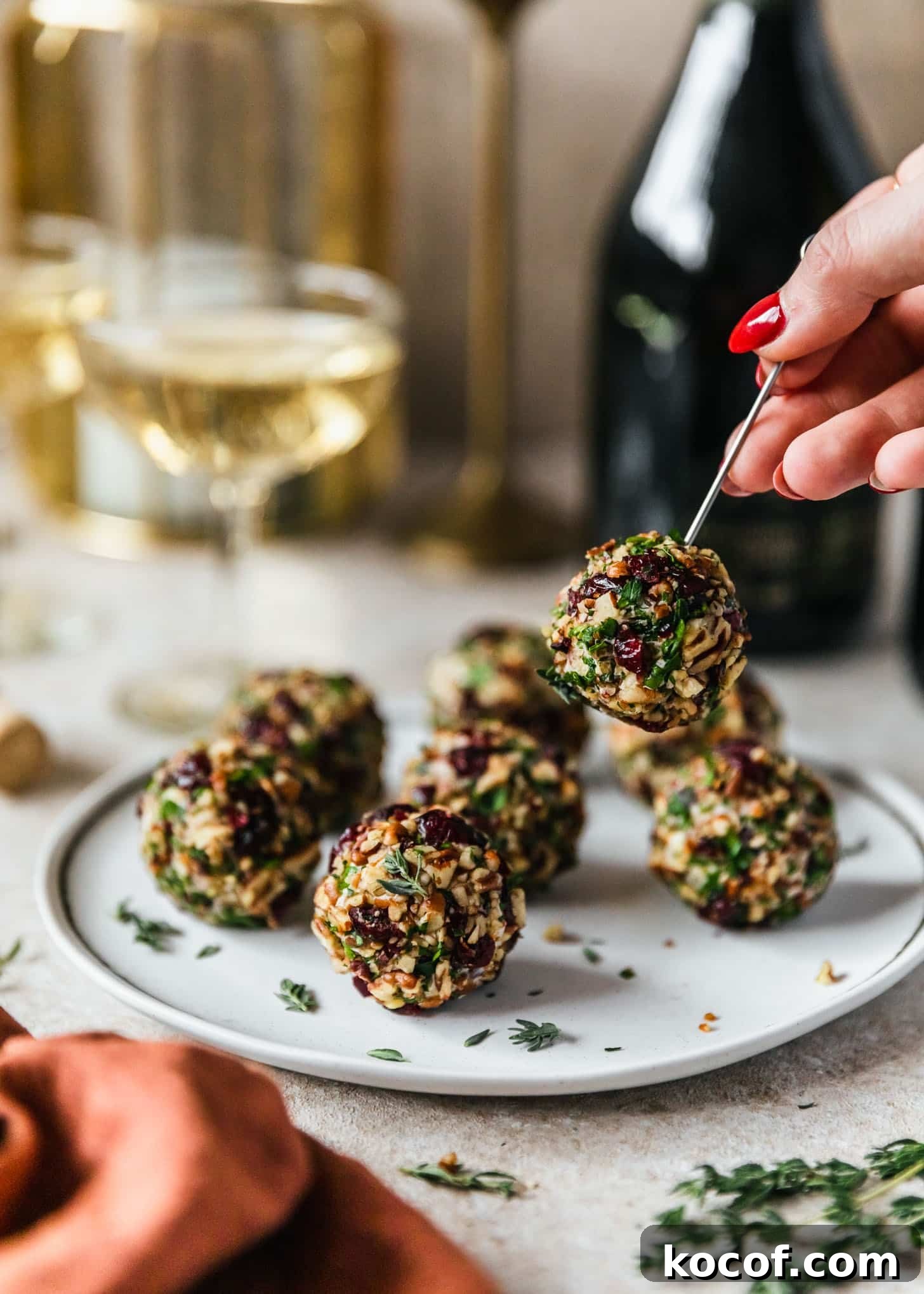 Cranberry Orange Cheesecake Bites: Festive Party Appetizer 10 A woman's hand with red nail polish grabbing a cranberry orange cheese ball off a plate next to champagne and an orange napkin on a tan table.