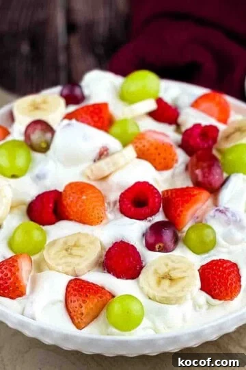 Creamy fruit salad in a white bowl, topped with fresh strawberries, raspberries, grapes, and bananas.