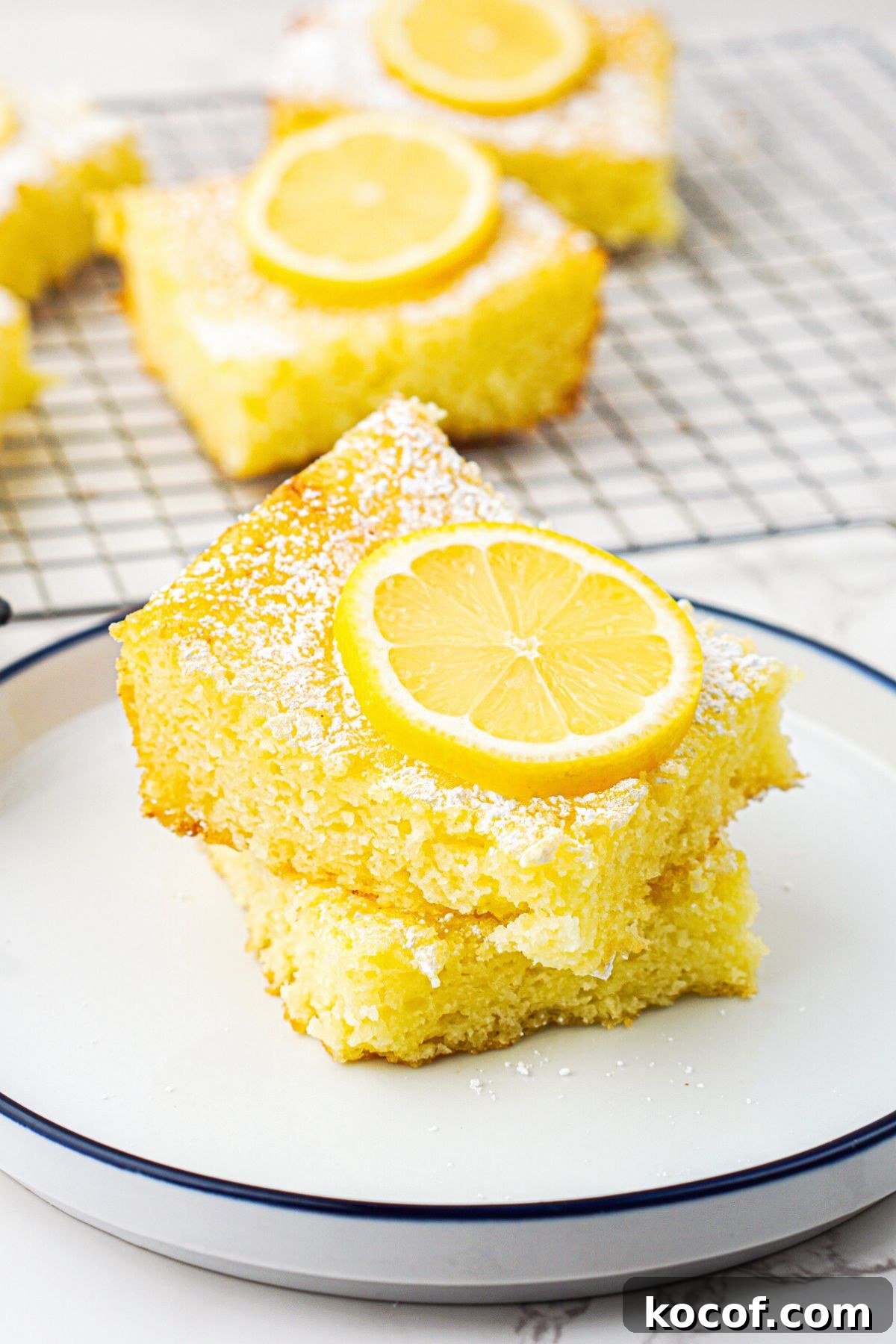 Bright yellow lemon bars stacked on a small white plate with a slice of lemon on top. 