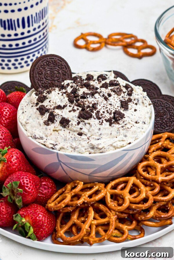 Strawberries, pretzels, and chocolate cookies, on a round white plate, around a cookies and cream Oreo dip, on a marble table.