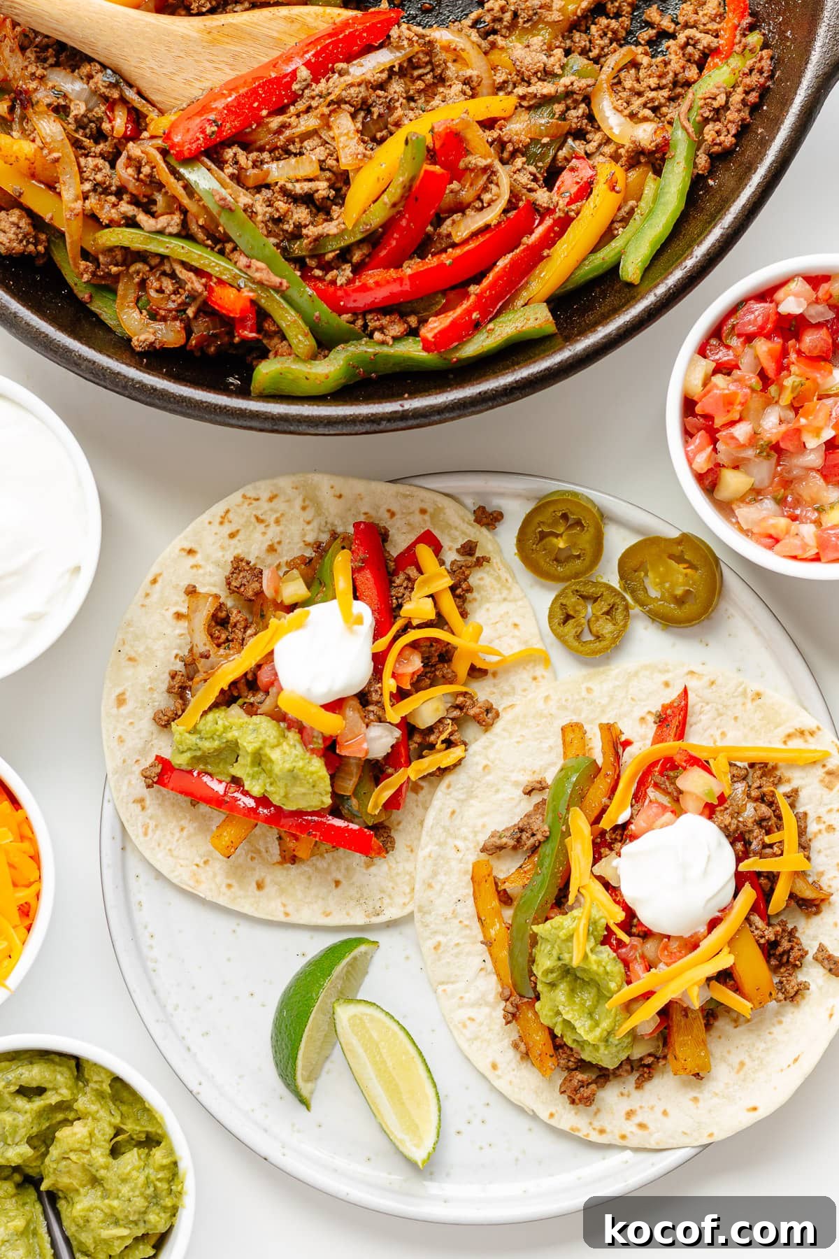 Quick Skillet Ground Beef Fajitas with Peppers and Onions 8 Two ground beef fajitas on flour tortillas on a plate with toppings and skillet of fajita mixture next to it.