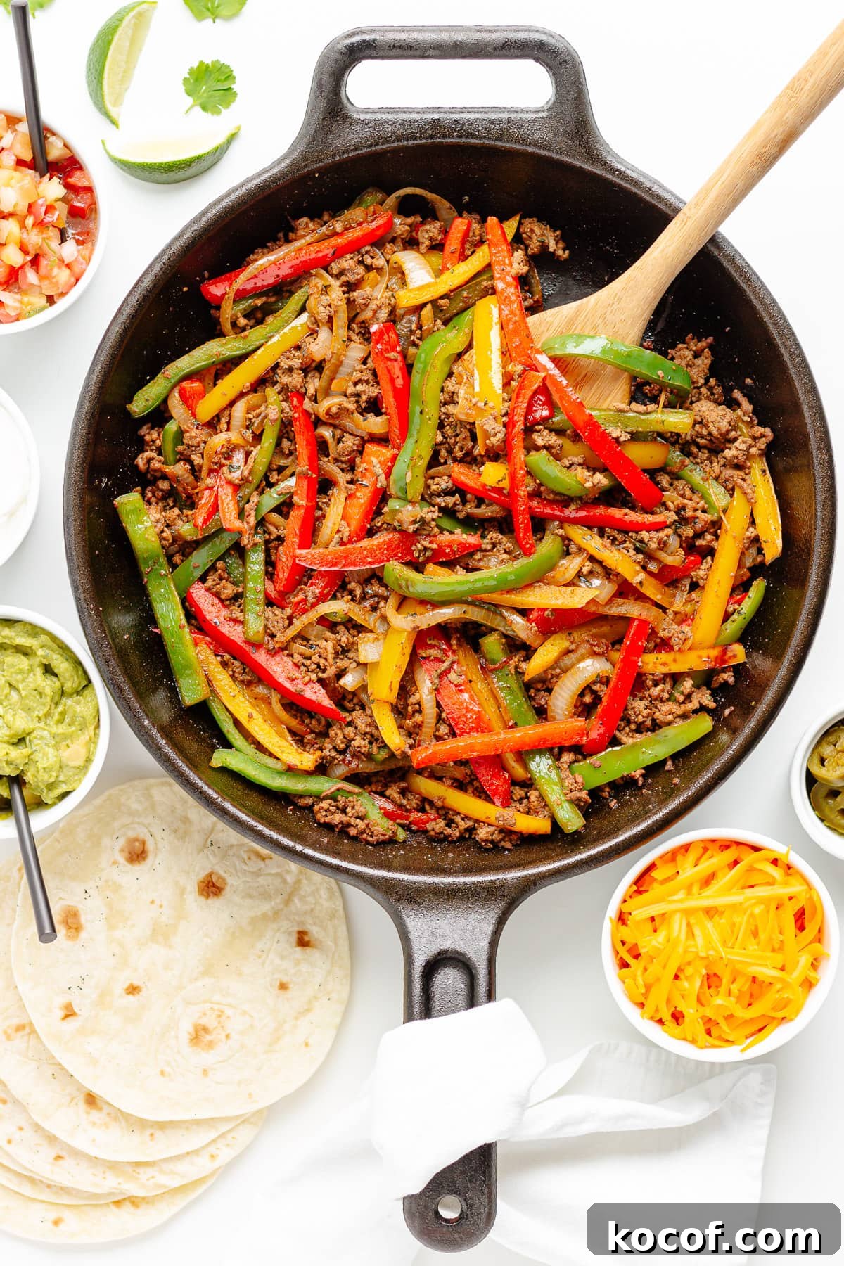 Quick Skillet Ground Beef Fajitas with Peppers and Onions 9 Ground beef fajitas filling in a cast iron skillet with wooden spoon, surrounded by a variety of toppings and tortillas.