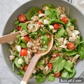 a large gray bowl filled with orzo, cherry tomatoes, spinach, mozzarella pearls, and chicken with two wooden serving spoons on a marble surface