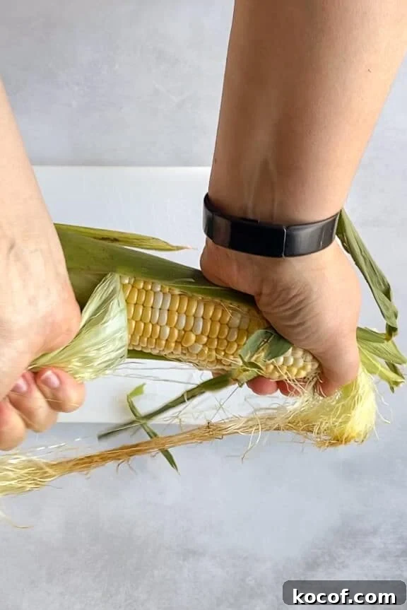 Hands are peeling the husk off a fresh ear of corn. The corn is yellow and surrounded by green leaves and silk.