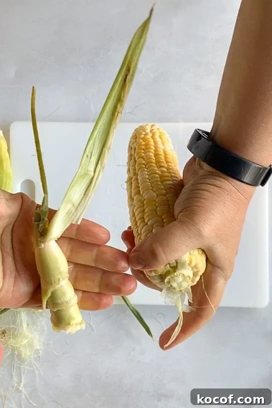 A pair of hands holding a corn cob and a broken stalk with husk.