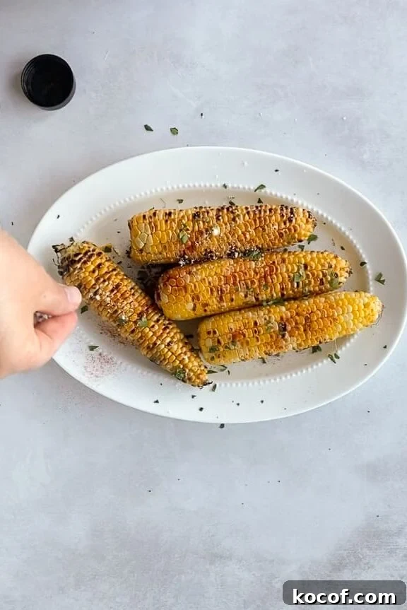 A hand sprinkles cheese over grilled corn on the cob on a white oval plate.