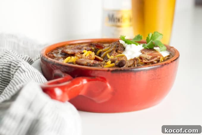 Smoked Brisket Chili Recipe: Bold, Smoky Chili with Beef Brisket 3 small red pot filled with brisket chili with cheese and sour cream and a garnish of cilantro, modelo beer bottle with glass of beer in background and napkins in foreground