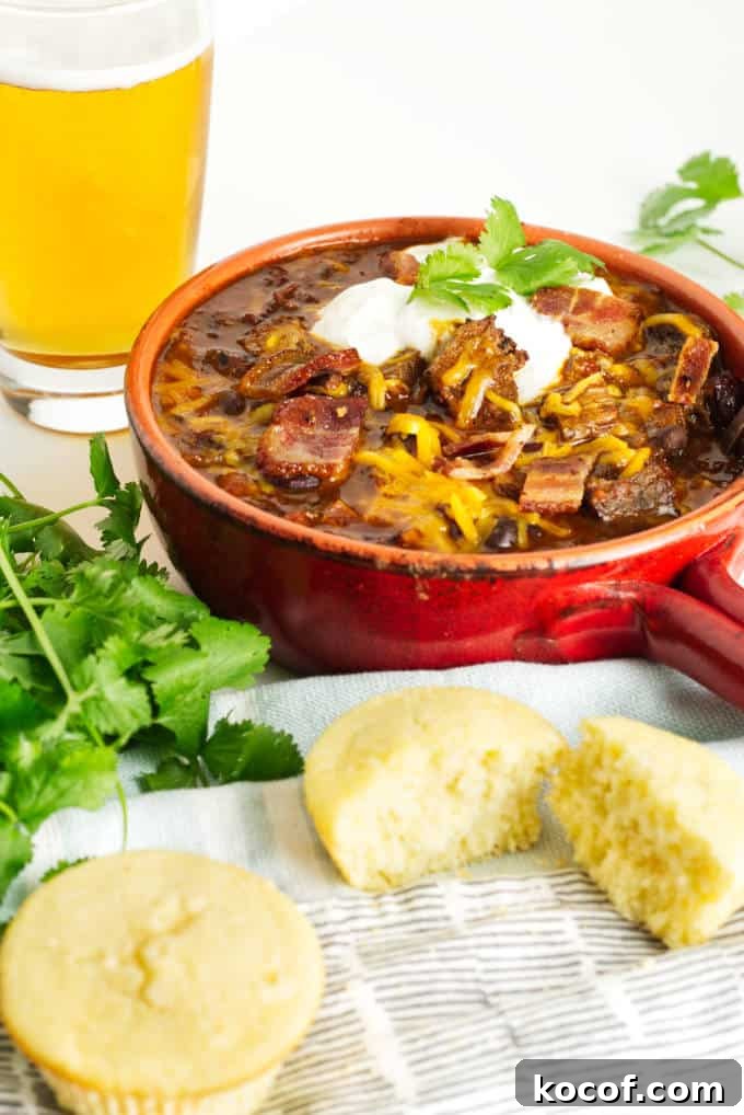 Smoked Brisket Chili Recipe: Bold, Smoky Chili with Beef Brisket 8 small red pot filled with brisket chili with cheese and sour cream and a garnish of cilantro, glass of beer in the background and cornbread in the foreground