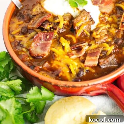 small red pot filled with brisket chili with cheese and sour cream and a garnish of cilantro, cornbread muffin in the foreground