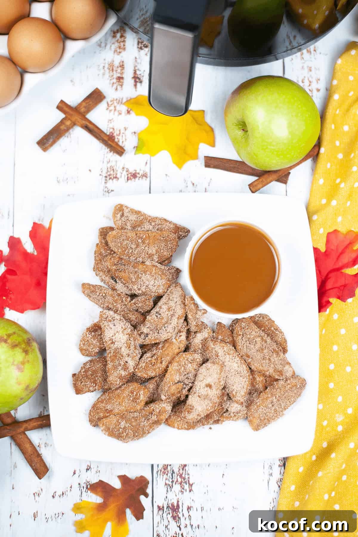 Crispy Pumpkin Spice Air‑Fried Apple Slices for Fall Snacks 6 A plate of air fried apples, cinnamon sugar-coated and served with a caramel dip. Surrounding items include eggs, green apples, cinnamon sticks, and autumn leaves on a wooden surface.