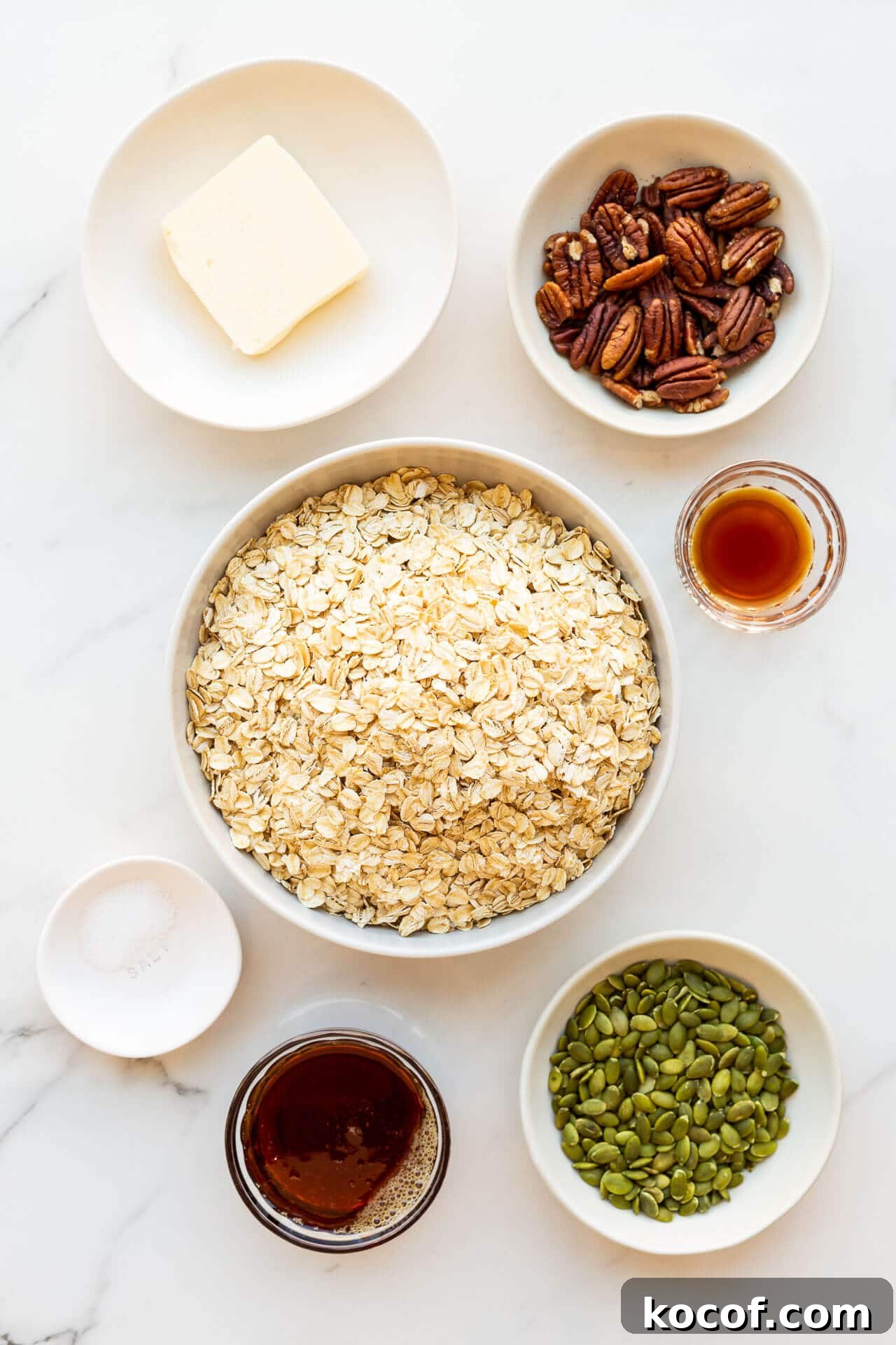Ingredients to make granola clusters measured out in bowls and ready to be mixed: butter, pecans, vanilla extract, oats, salt, maple syrup, and pumpkin seeds