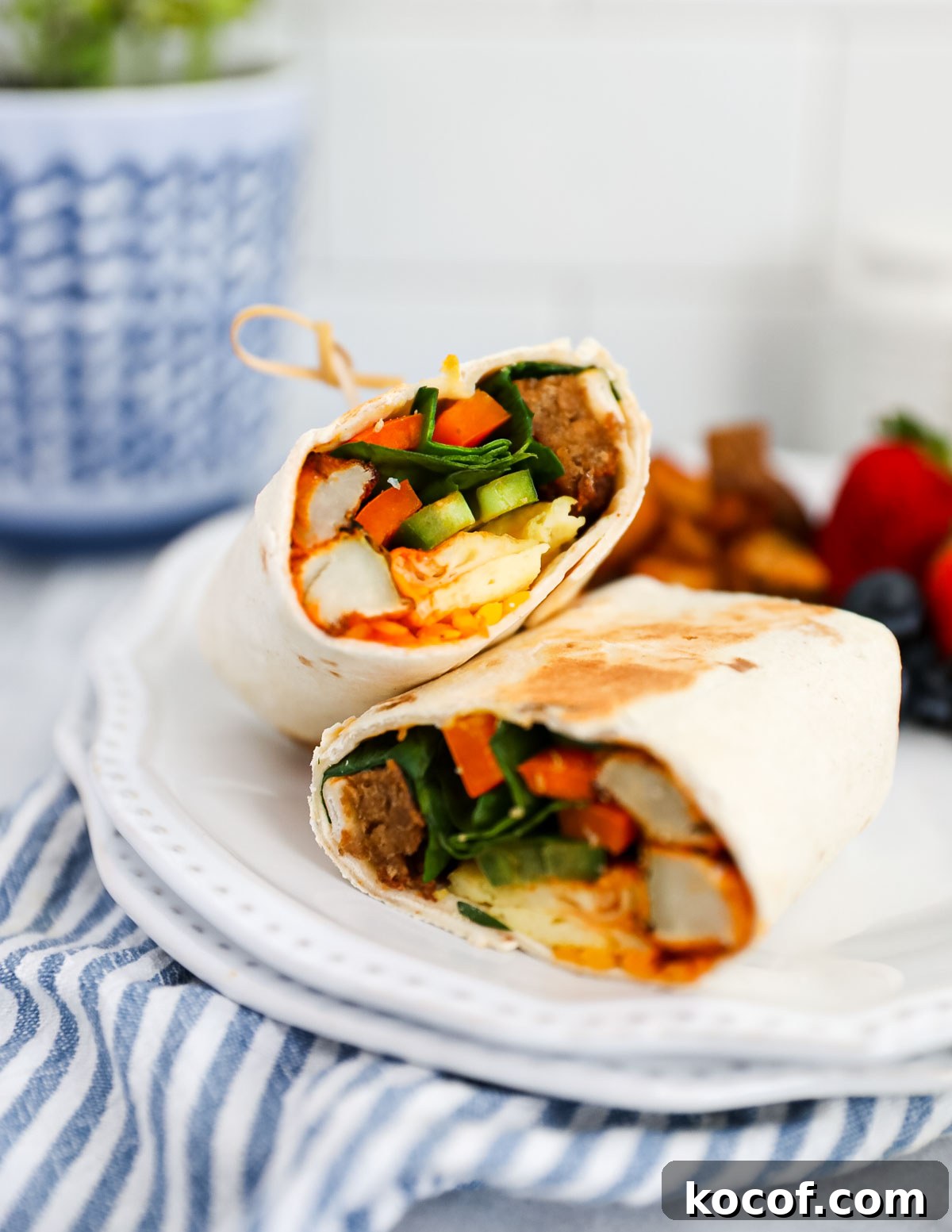 A plant based breakfast burrito filled with plant based egg and sausage, fresh vegetables, and breakfast potatoes. It is cut in half and on a plate with berries