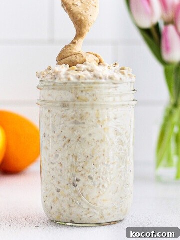A picture of a clear mason jar with overnight oatmeal inside. A spoon is drizzling peanut butter on top. There are pink tulips and a sliced orange in the background.