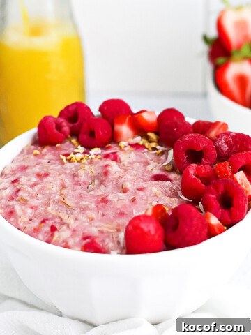 Pink raspberry oatmeal in a white bowl, garnished with fresh raspberries and strawberries. There is a bowl of strawberries and orange juice in the background.