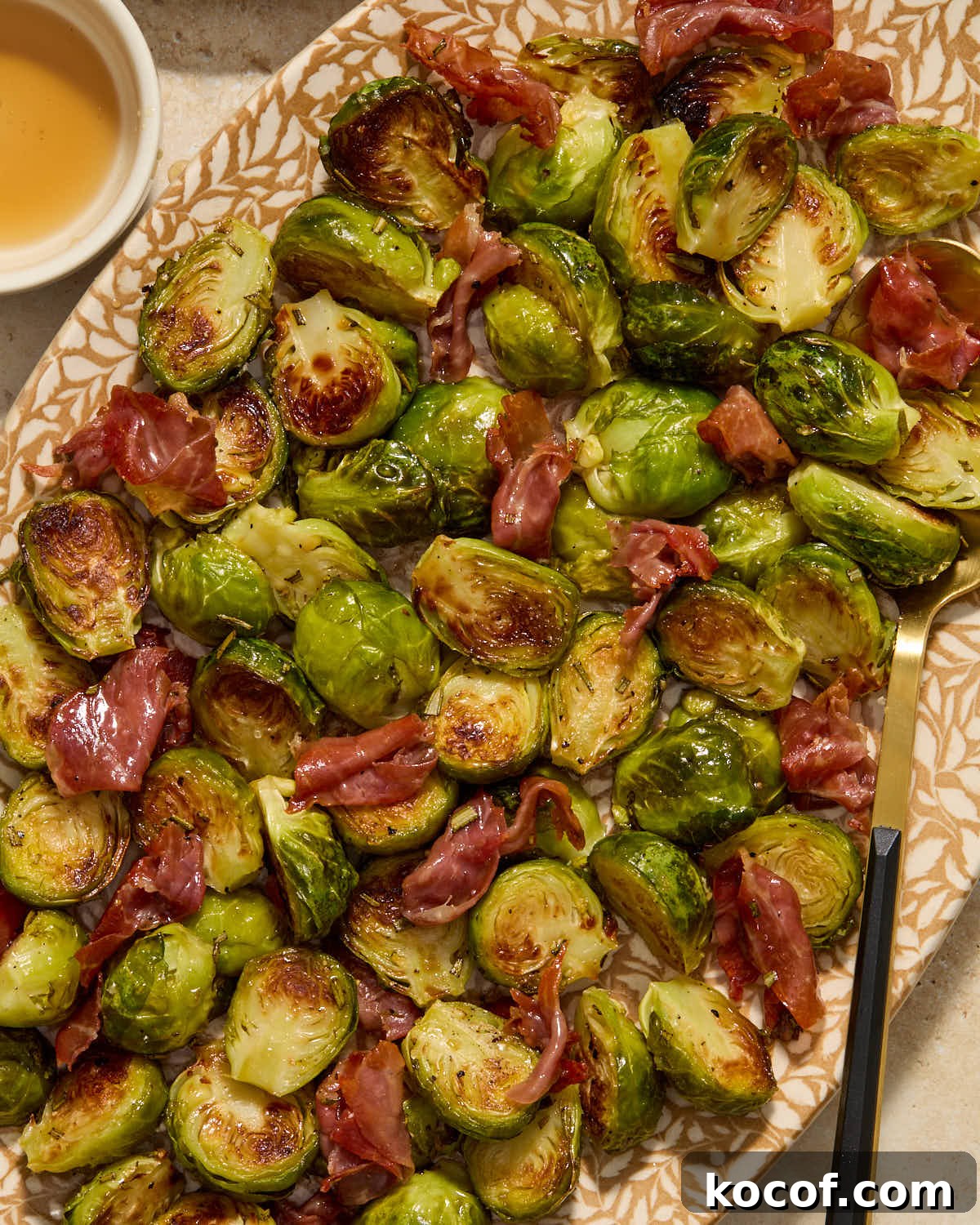 Brussels sprouts and prosciutto served on a platter with a serving spoon and maple syrup on the side.