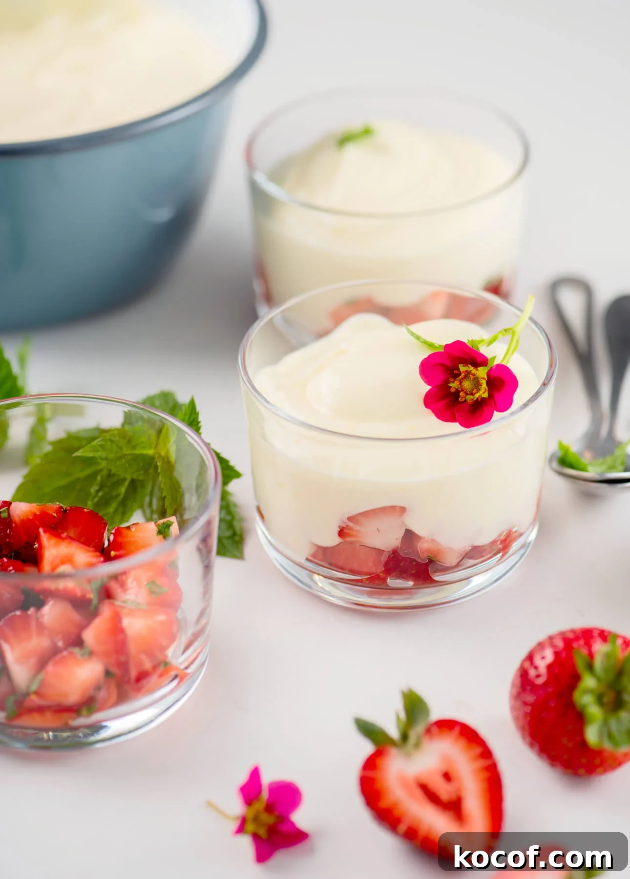 Lemon-Lime Citrus Mousse with Macerated Strawberries 2 Three glass cups with layered dessert of chopped strawberries and citrus mousse, garnished with a small pink flower and mint. Fresh strawberries and mint leaves are scattered on a white surface nearby. // FoodNouveau.com