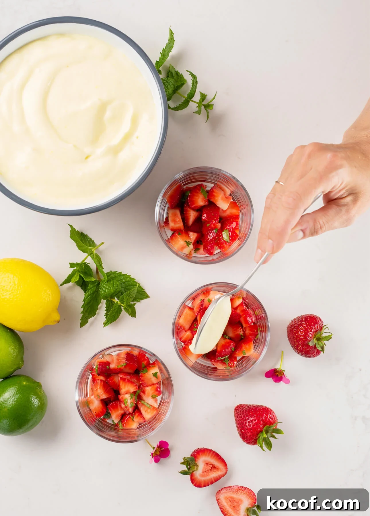 Lemon-Lime Citrus Mousse with Macerated Strawberries 4 A hand spoons creamy citrus mousse over diced strawberries in glass cups, surrounded by whole strawberries, lemon, limes, mint leaves, and a bowl of cream on a white surface. // FoodNouveau.com