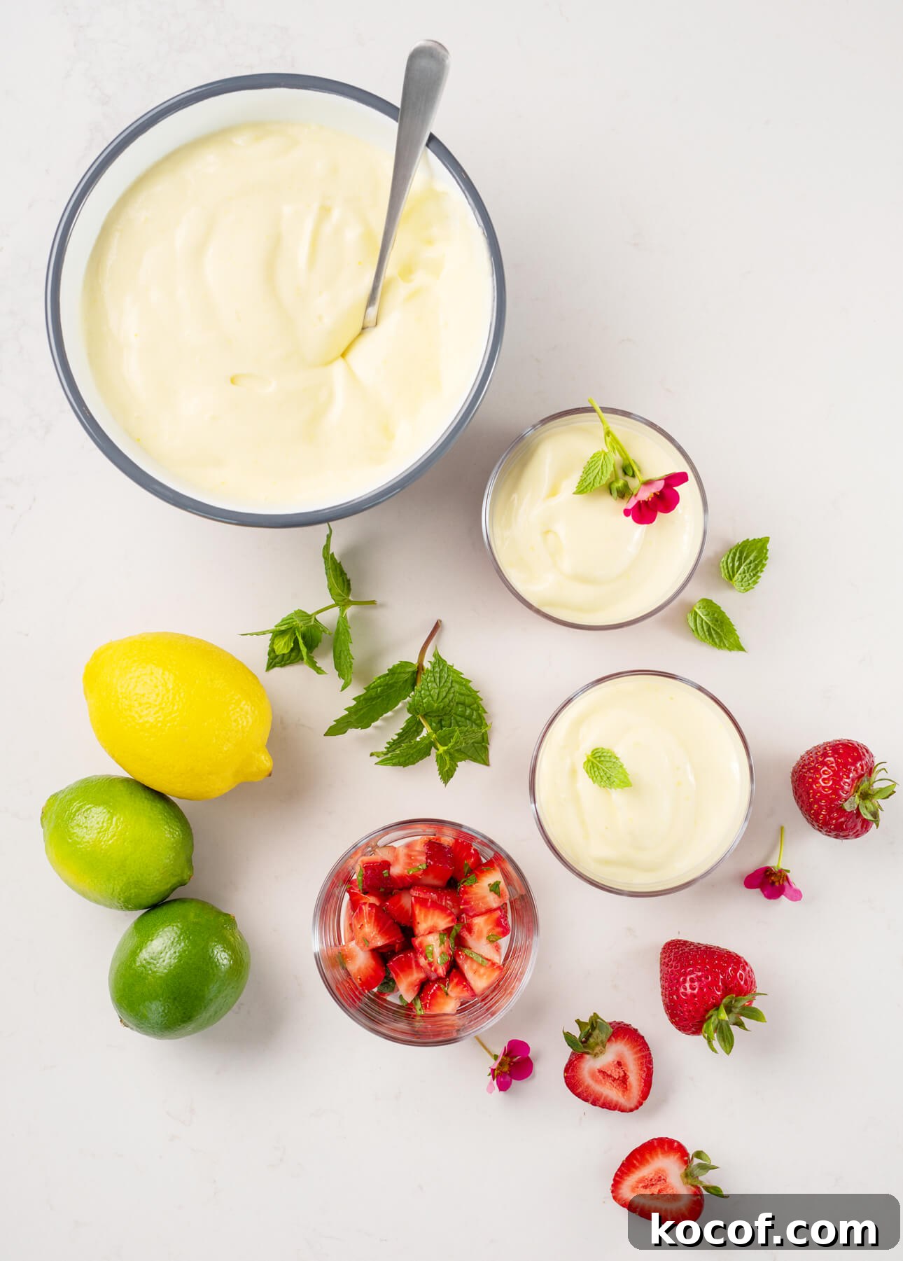 Lemon-Lime Citrus Mousse with Macerated Strawberries 6 A bowl and small glasses of creamy citrus mousse are arranged with whole lemons, limes, mint leaves, strawberries, a dish of diced strawberries, and small pink flowers on a white surface. // FoodNouveau.com