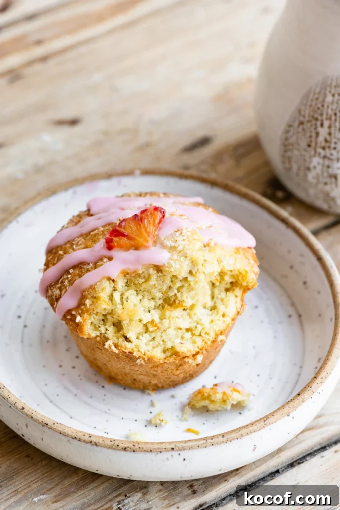 A single orange coconut muffin topped with an orange vanilla glaze and a small orange slice on a plate, with a bite missing.