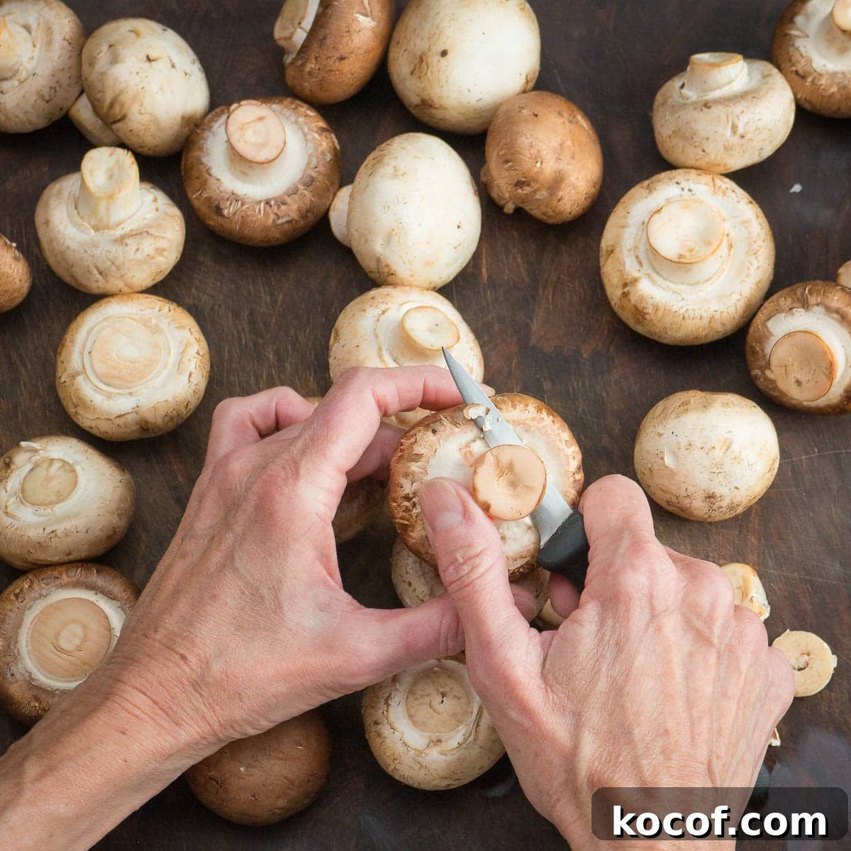 Hands trimming mushrooms with a paring knife on a walnut cutting board.