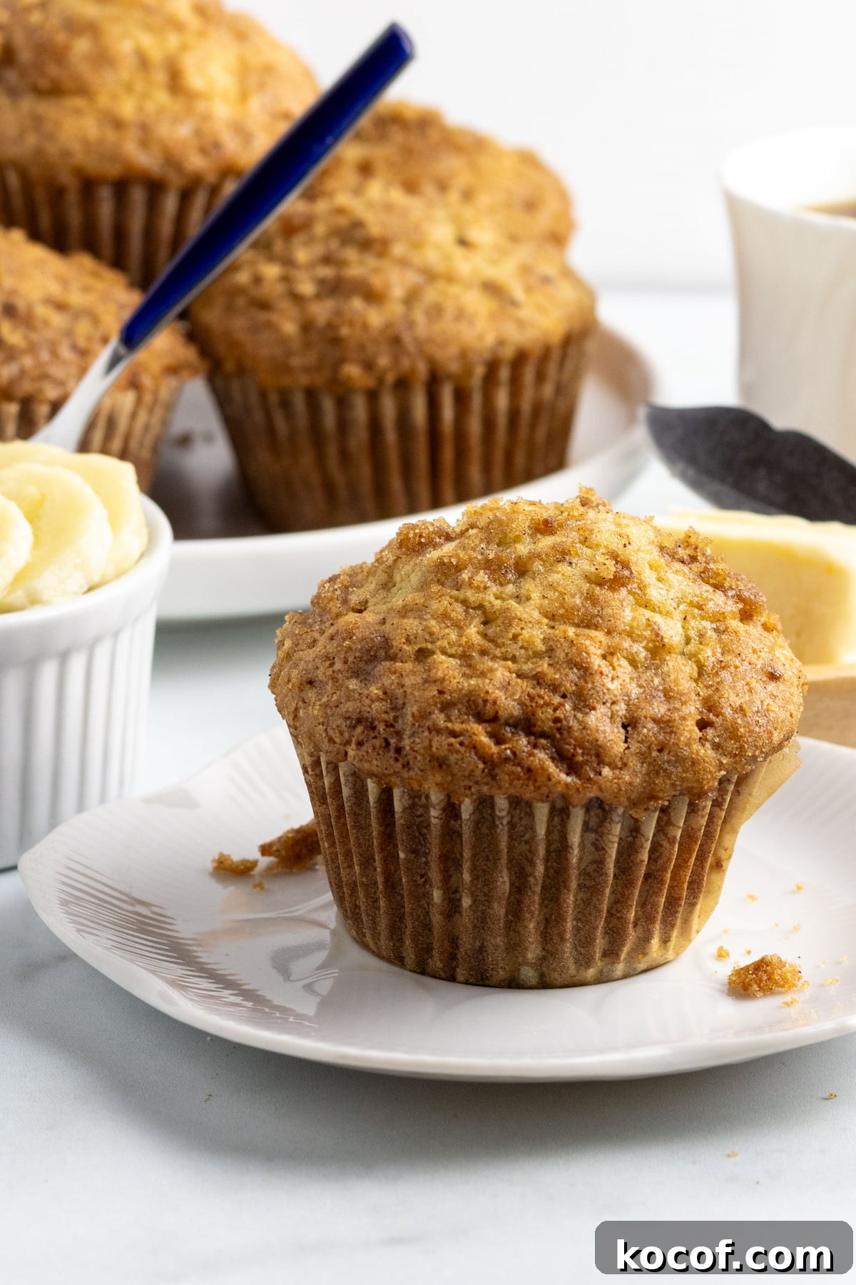 Moist Banana Muffins Without Dairy — Simple Vegan Recipe 3 dairy free banana muffin on a small white plate with a plate of muffins, a cup of fruit and a cup of coffee behind it