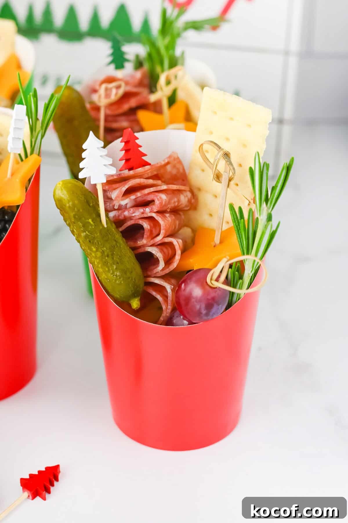Christmas charcuterie cups - red paper cup filled with charcuterie ingredients: folded salami skewers, cheddar cheese stars, a gherkin, a grape, a cracker, and rosemary. 