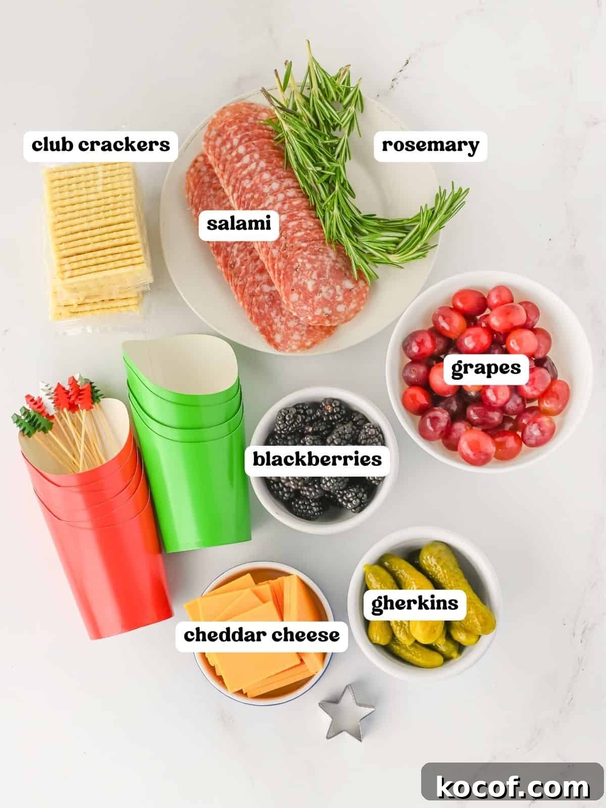 : Flat lay image showing labeled ingredients for charcuterie cups. Items include club crackers, rosemary sprigs, sliced salami on a plate, red grapes in a bowl, blackberries in a bowl, cheddar cheese slices in a bowl, and gherkins in a bowl. Red and green paper cups and decorative skewers are also visible.