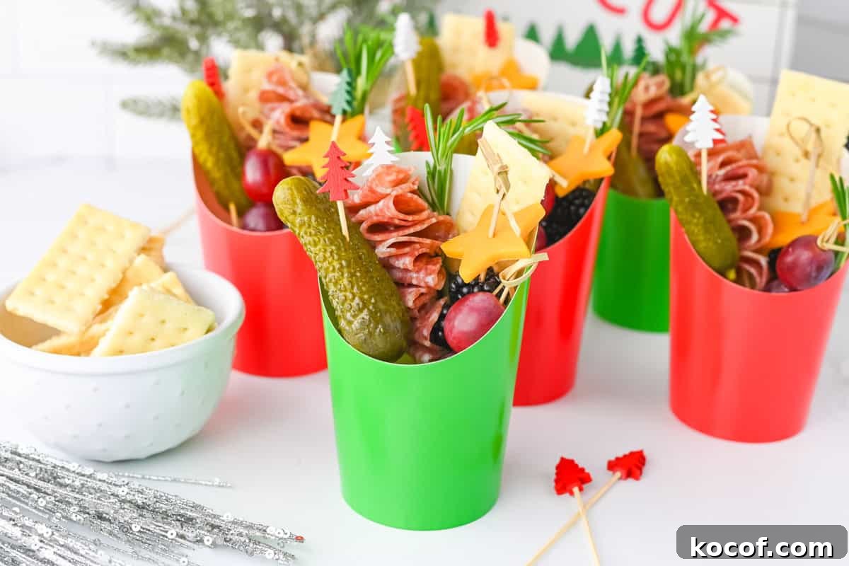 Red and green paper charcuterie cups are displayed together, each filled with folded salami skewers, cheddar cheese stars, grapes, gherkins, crackers, and rosemary.