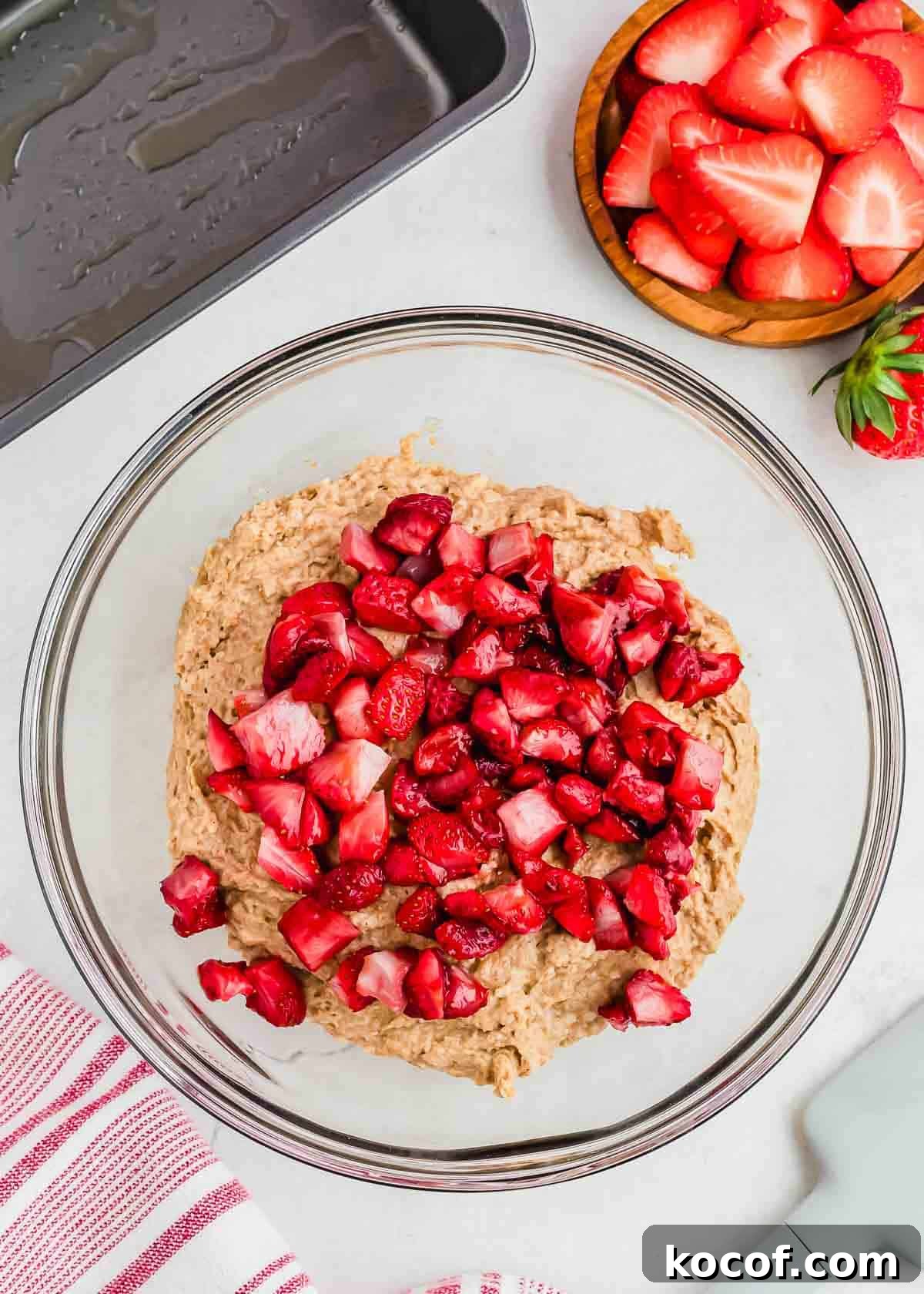A glass bowl contains bread batter topped with chopped roasted strawberries, ready to be mixed. Nearby, a small wooden bowl holds more sliced strawberries. A red and white striped cloth and a baking tray are visible in the background.